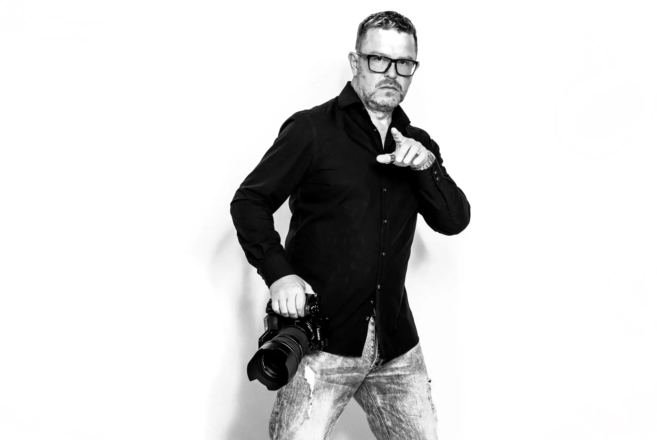 Jens Achtert in Schwarzweiß mit Kamera in der Hand, zeigt in die Kamera, vor weißem Hintergrund · Businessfotograf aus Calw bei Stuttgart zu Datenlieferung, Dateiformaten und Farbräumen