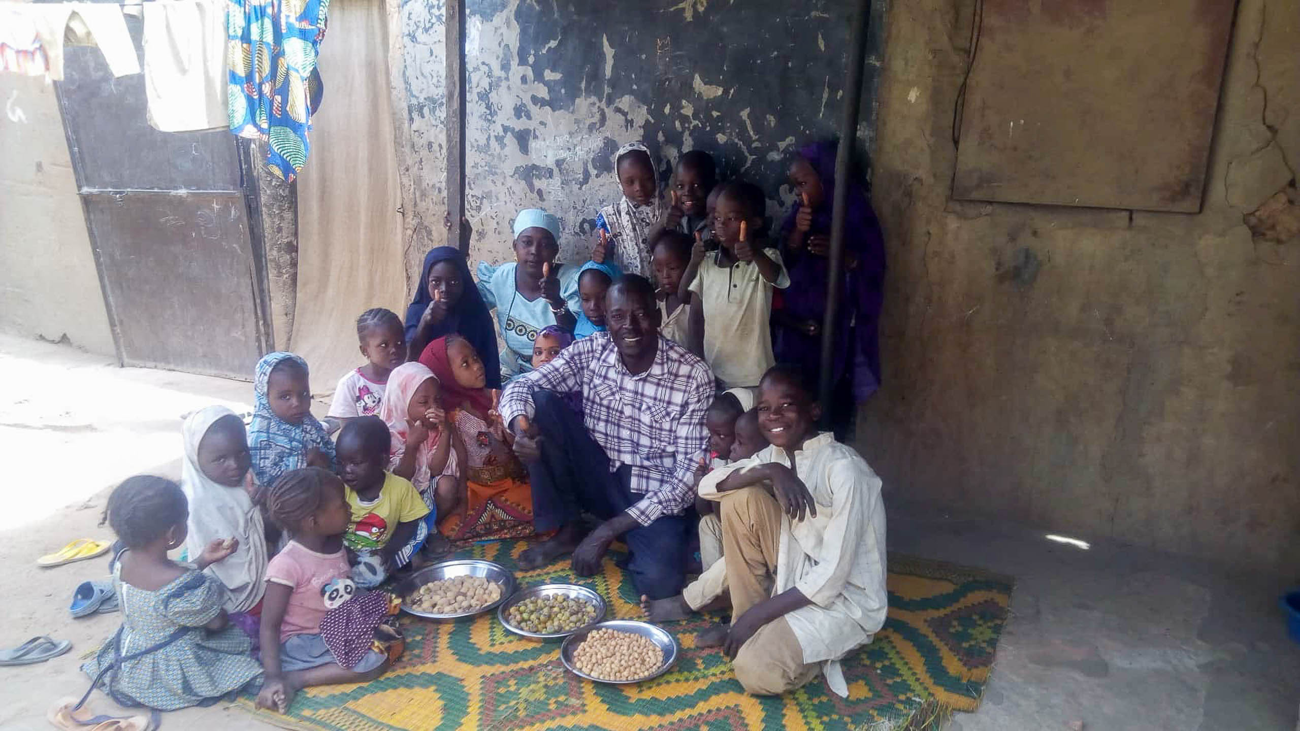 Mourtala Issa sitzt mit vielen Kindern seiner Familie und Nachbarschaft vor einer Lehmwand in Zinder, Niger · Projekt #waterforzinder